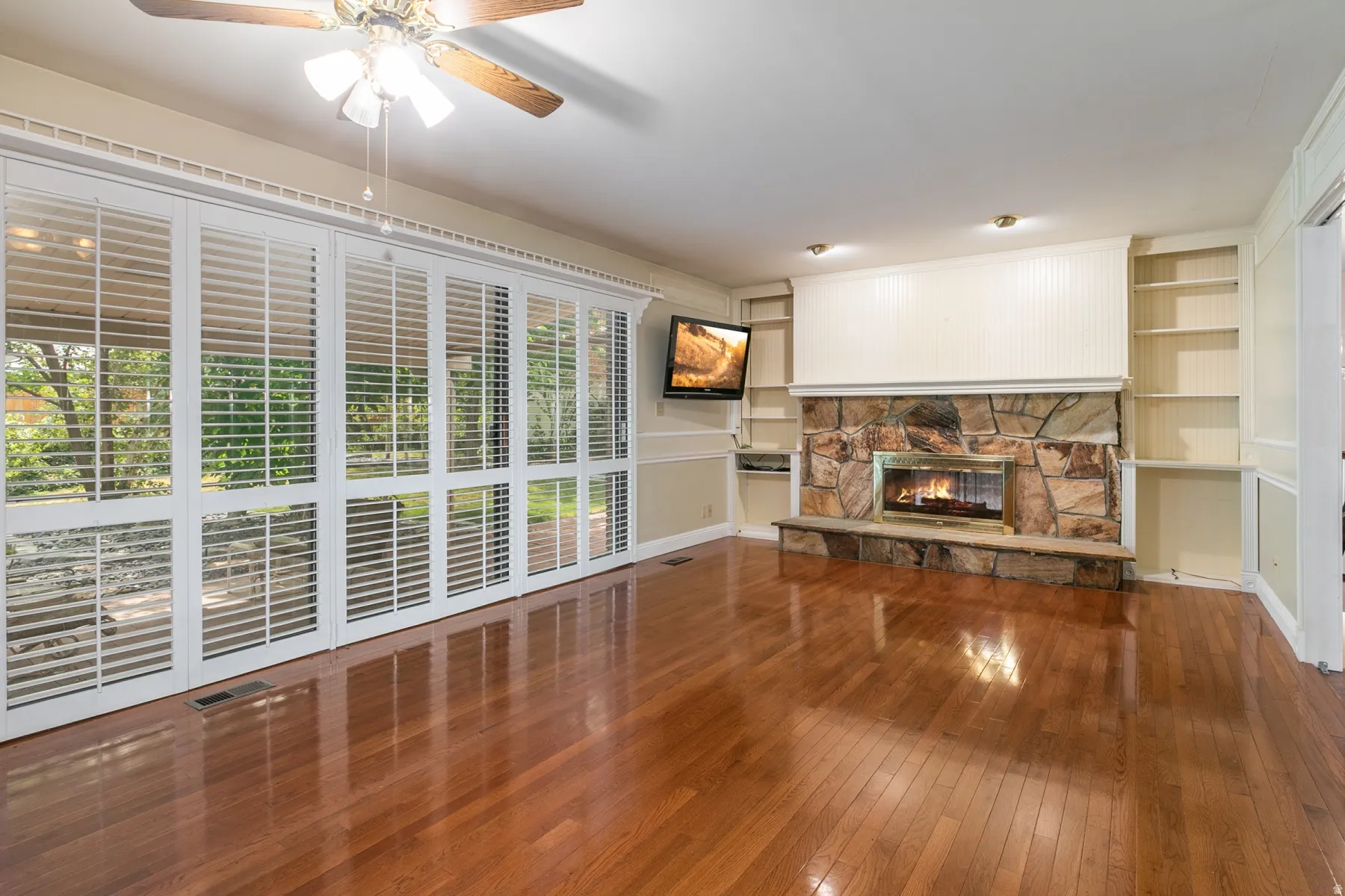Stone-clad fireplace with built-in shelving, wood-finish flooring, and large windows with plantation shutters