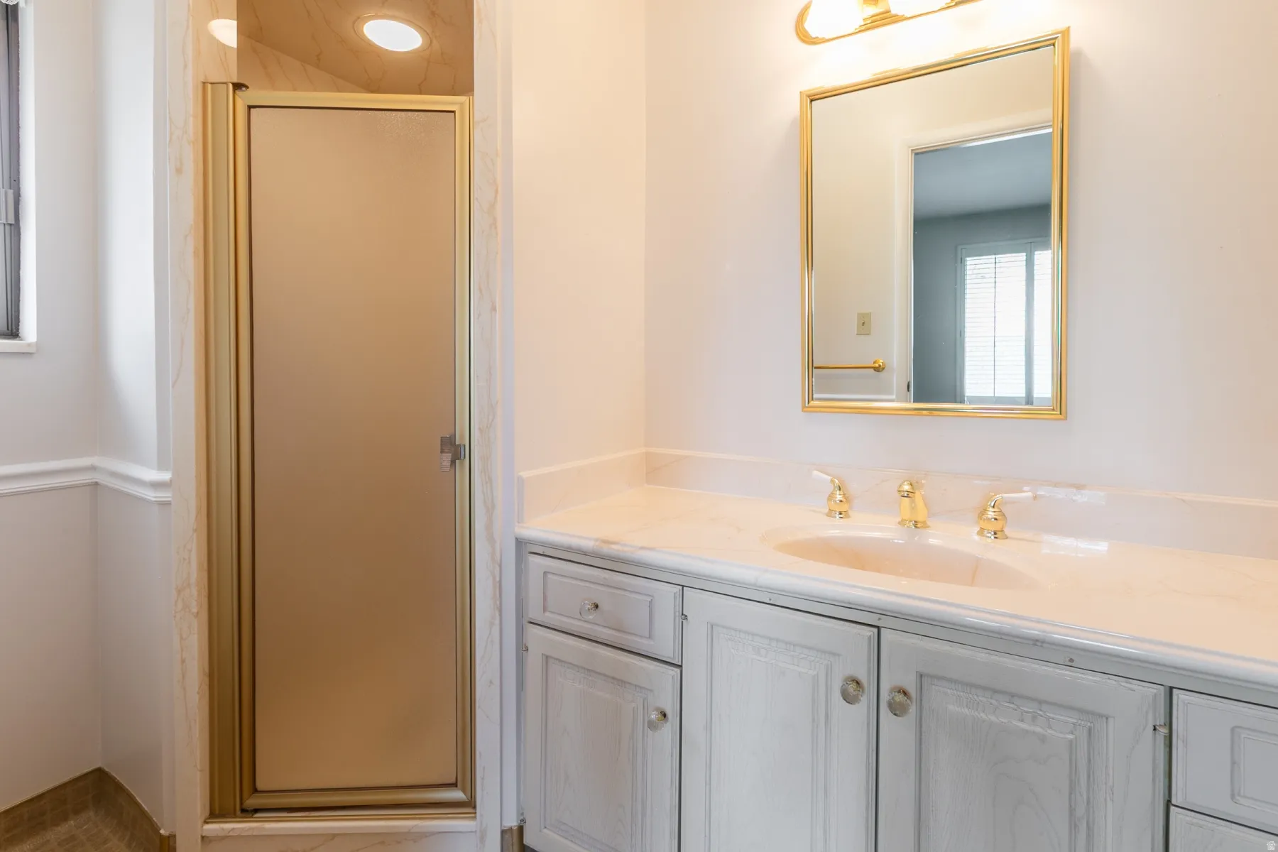 Vanity with integrated sink and gold-tone fixtures, wall-mounted mirror with gold-tone frame, painted cabinetry, and a shower enclosure with a frosted glass door and gold-tone frame