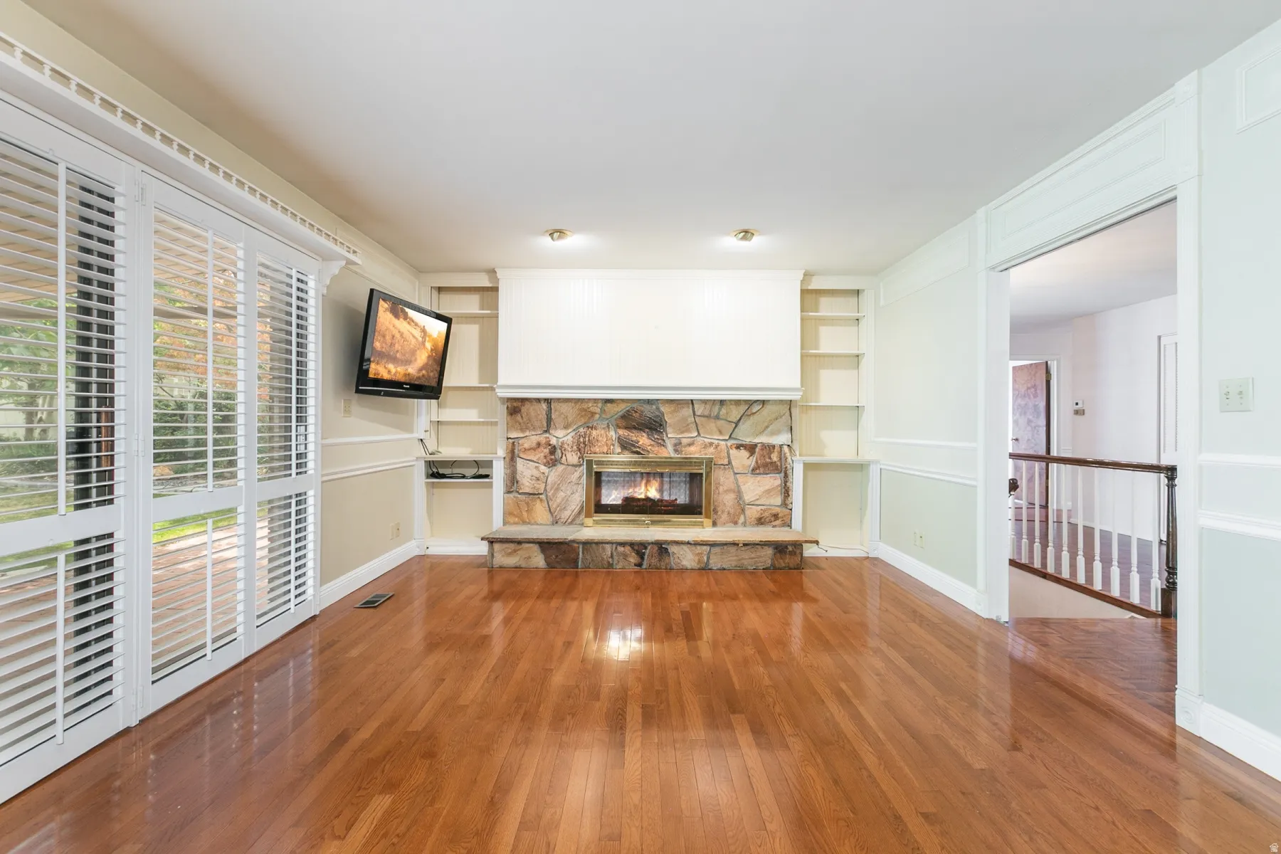 Hardwood flooring, stone fireplace with hearth, built-in shelving, crown molding, and plantation shutters