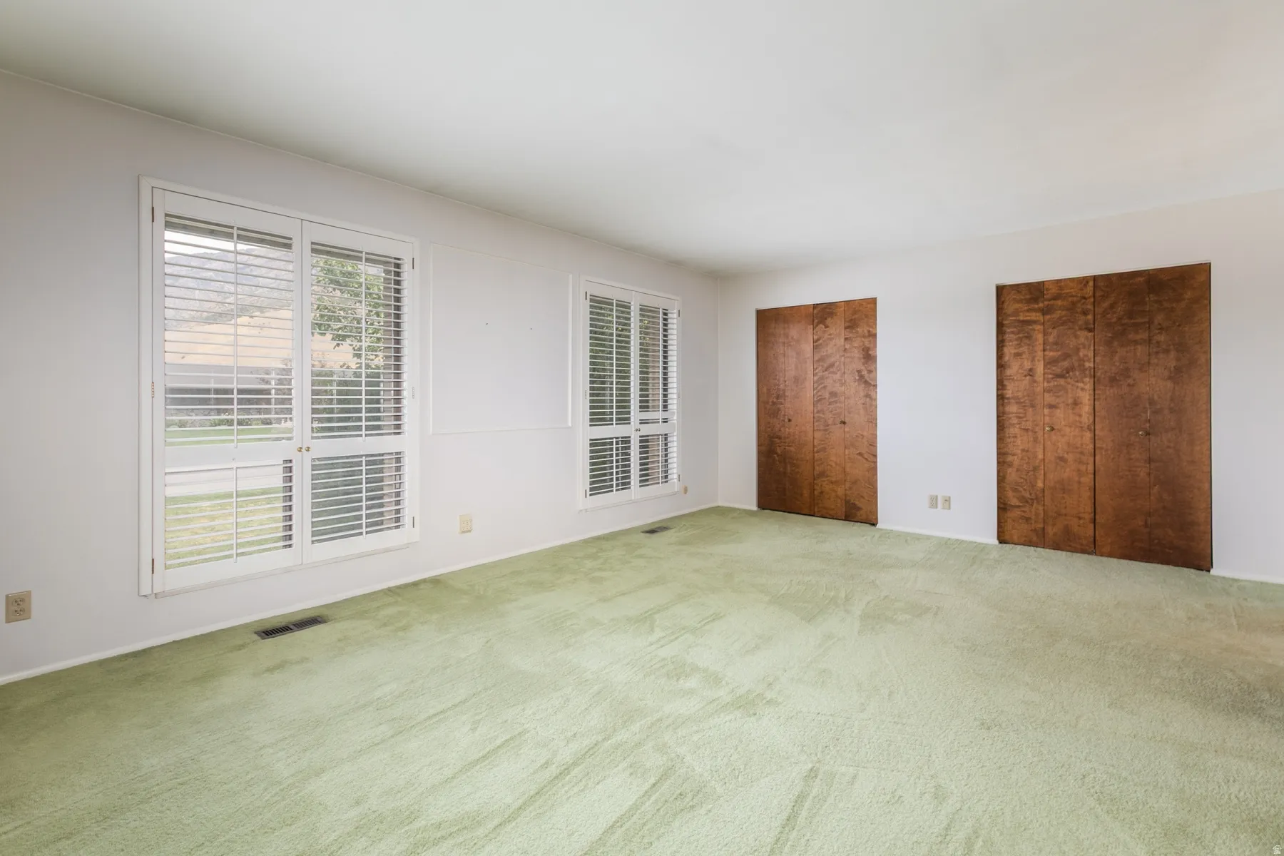 Spacious room featuring dual windows with white plantation shutters and two sets of wood-paneled bi-fold closet doors