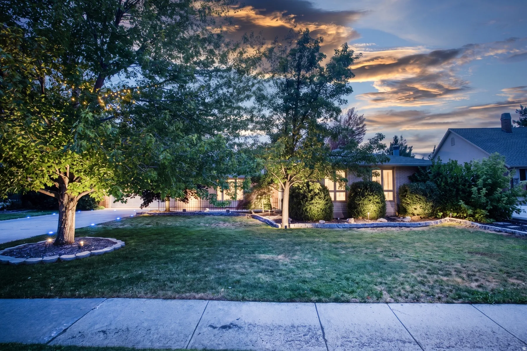 Manicured front lawn with mature trees, established landscaping, decorative lighting, and a concrete sidewalk