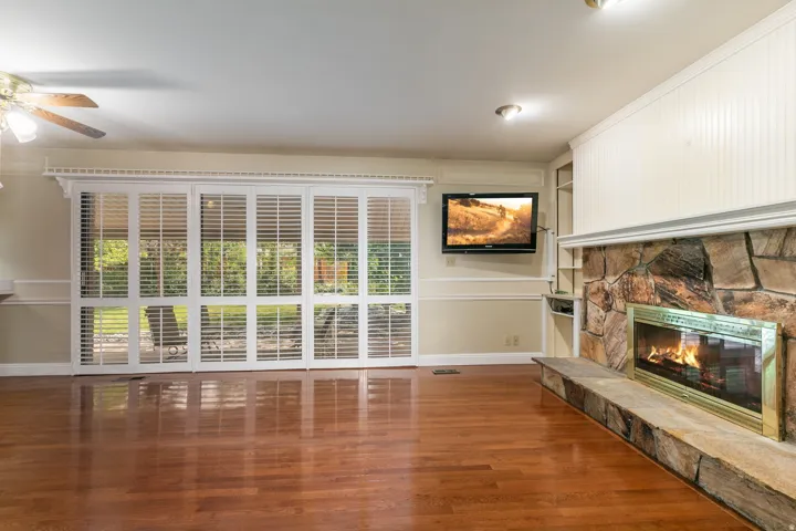 Spacious living area featuring rich wood-finish flooring, white plantation shutters across expansive windows, ceiling fan with recessed lighting, wall-mounted flat-screen television, and a decorative stone fireplace with built-in mantel