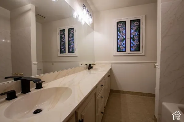 Double vanity featuring stone-finish countertops, matte black widespread faucets, floor-to-ceiling mirror, and dual windows with decorative leaded glass inserts