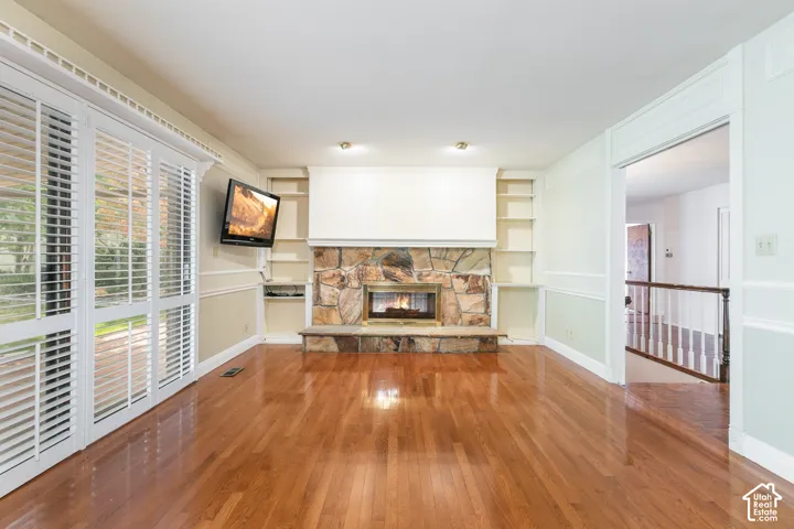 Hardwood flooring, stone fireplace with hearth, built-in shelving, crown molding, and plantation shutters