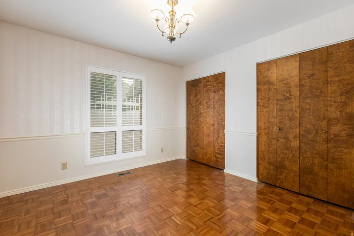 Parquet wood flooring, a window with plantation shutters, two paneled closet doors, and an overhead chandelier