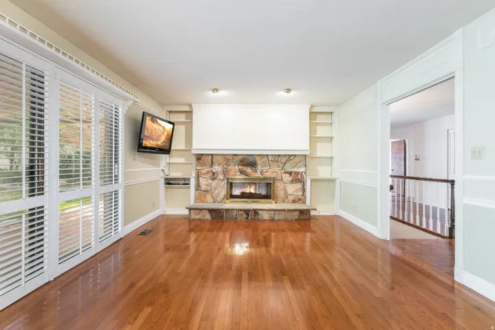 Hardwood flooring, stone fireplace with hearth, built-in shelving, crown molding, and plantation shutters