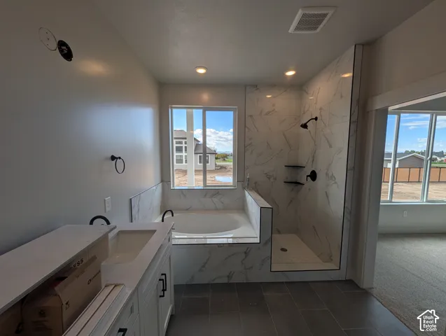 Bathroom featuring a deep soaking tub, walk-in shower with tile surround, dual-sink vanity with white countertop, and dark-toned floor tiling