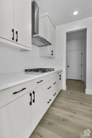 White cabinetry with matte black hardware, stainless steel range hood, built-in gas cooktop, light-toned wood-finish flooring, and white countertops