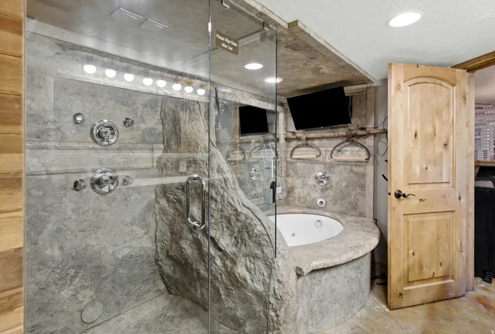 Glass-enclosed shower with multiple sprayers, integrated jetted tub, and stone-finish wall surrounds