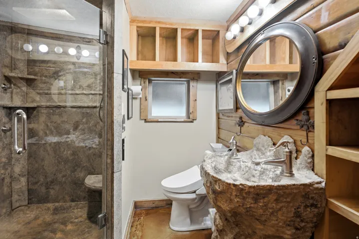 Stone-look shower with glass enclosure, custom rock-finish vanity with dual sinks, wall-mounted light fixture, and wood paneling