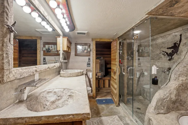 Stone vanity with integrated basin sinks, glass-enclosed shower, wood-paneled walls, and a built-in sauna