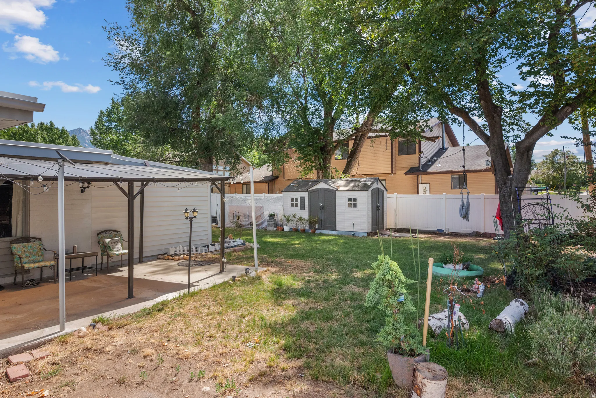 Expansive backyard featuring a covered patio, concrete hardscaping, a white privacy fence, mature trees, and two storage sheds