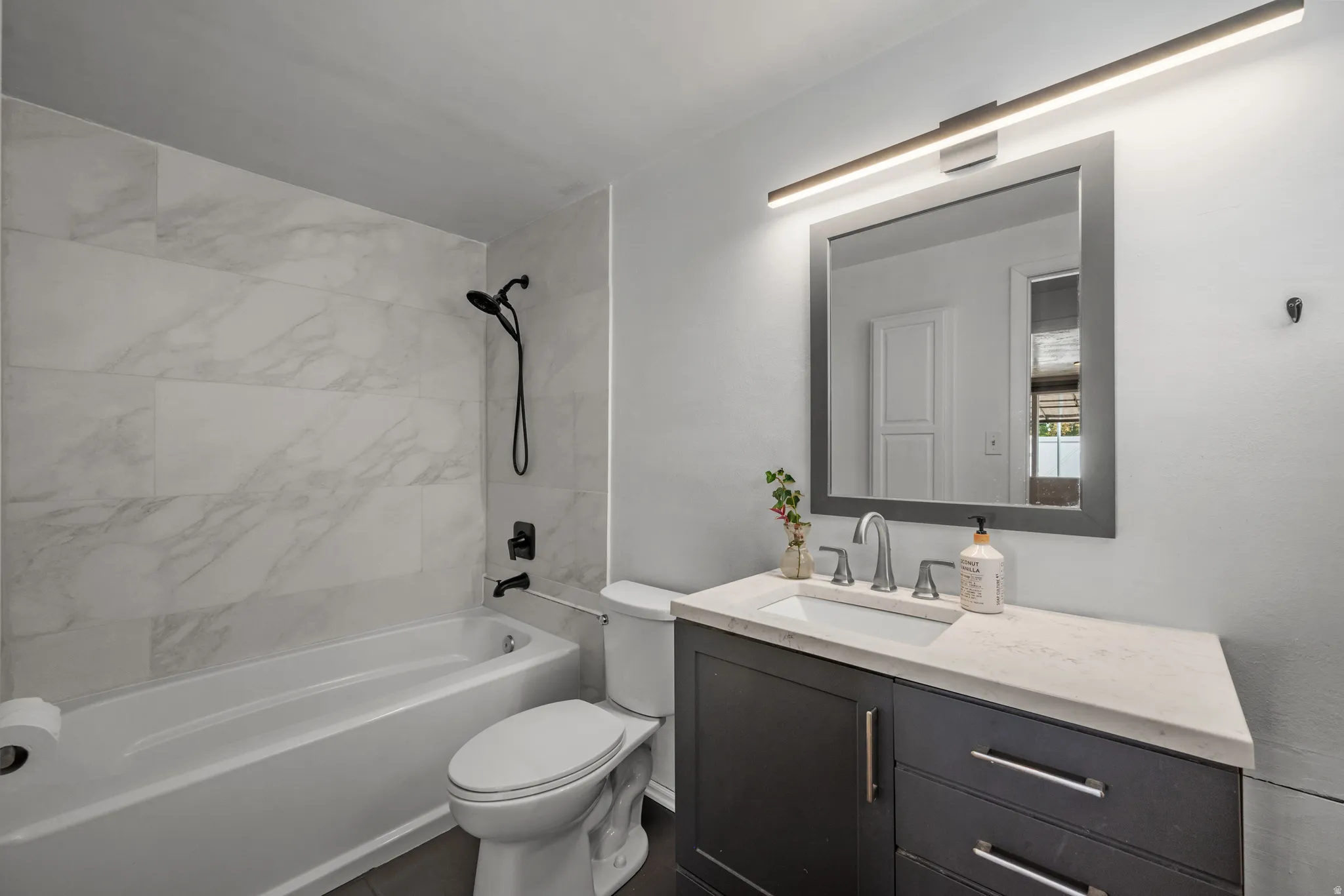 Bathroom featuring a white bathtub, black shower fixtures, tiled shower surround, dark vanity with an integrated sink, and a rectangular mirror with overhead lighting