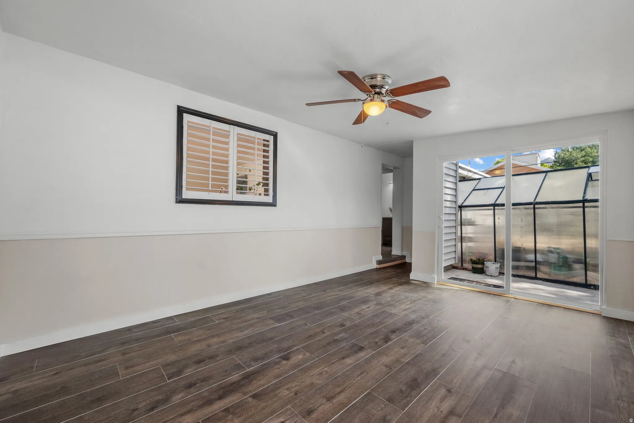 Wood-finish flooring, two-tone wall treatment, ceiling fan with light fixture, window with plantation shutters, and sliding glass door