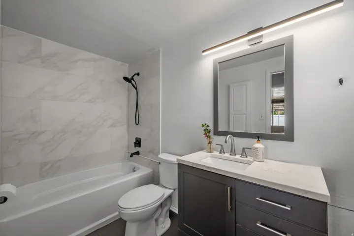 Bathroom featuring a white bathtub, black shower fixtures, tiled shower surround, dark vanity with an integrated sink, and a rectangular mirror with overhead lighting