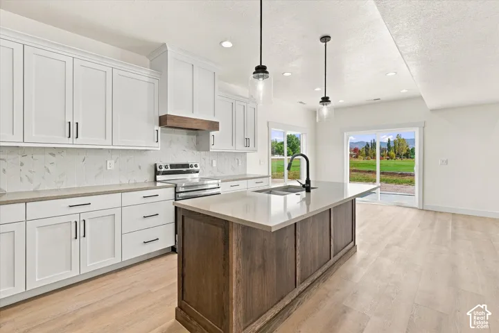 White shaker cabinetry, light wood-finish flooring, a kitchen island with sink and pendant lighting, stainless steel range, and a sliding glass door to exterior