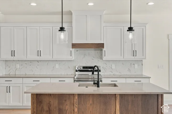 Kitchen featuring white shaker cabinetry, patterned tile backsplash, stainless steel range, light-toned countertops, and a wood-finish island with an undermount sink