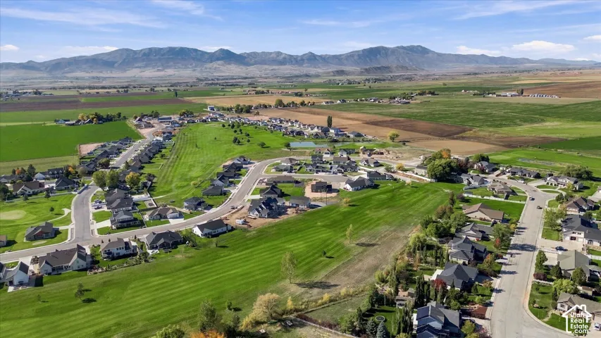 Residential community with paved streets, surrounded by expansive green spaces and agricultural fields, with distant mountains forming a natural backdrop