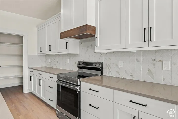 White shaker cabinetry, wood-finish range hood, stainless steel electric range, herringbone pattern tile backsplash, and light-toned countertops