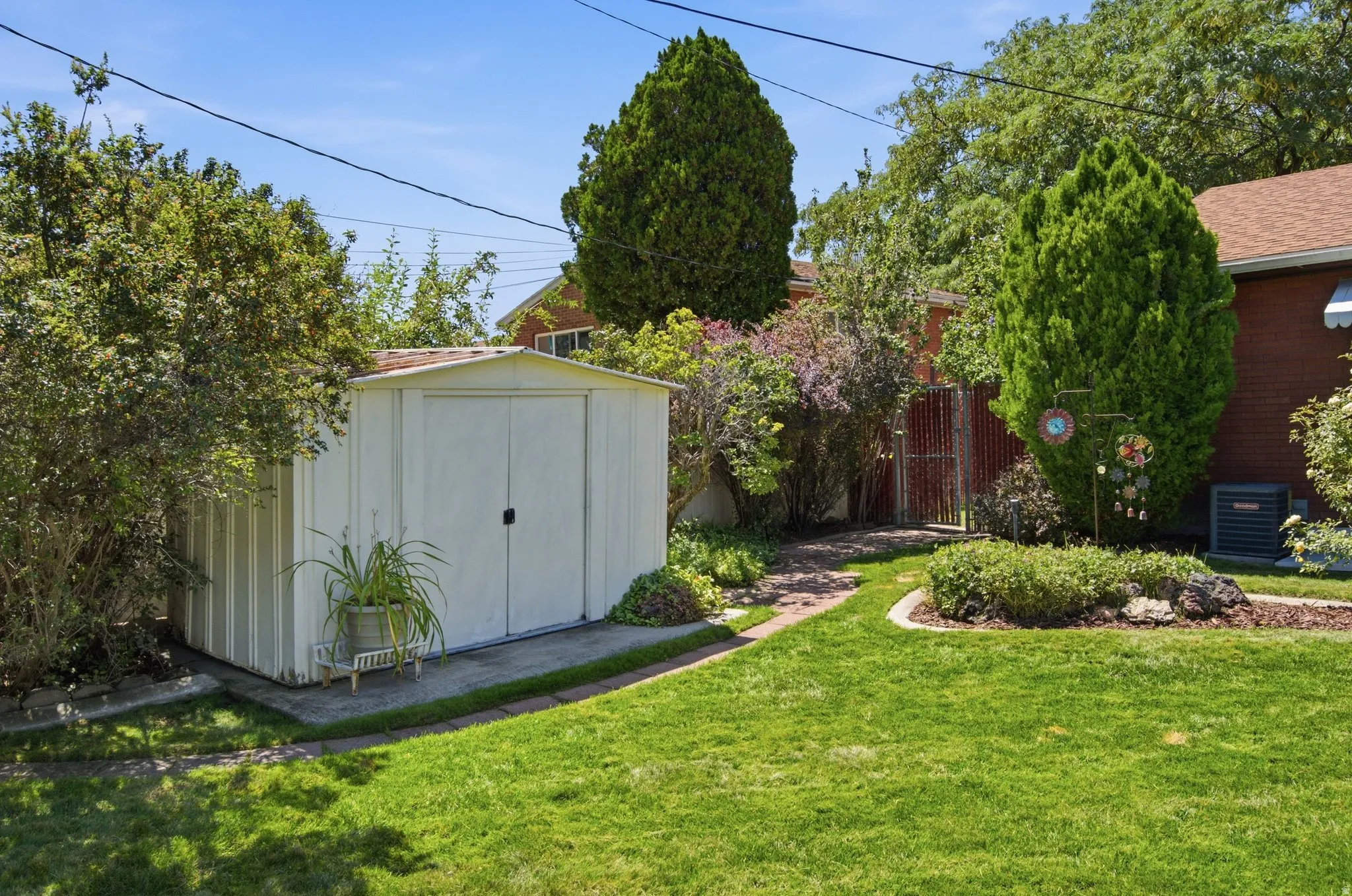 Spacious backyard featuring a manicured lawn, mature landscaping with evergreen trees and flowering shrubs, and a detached storage shed with white metal siding