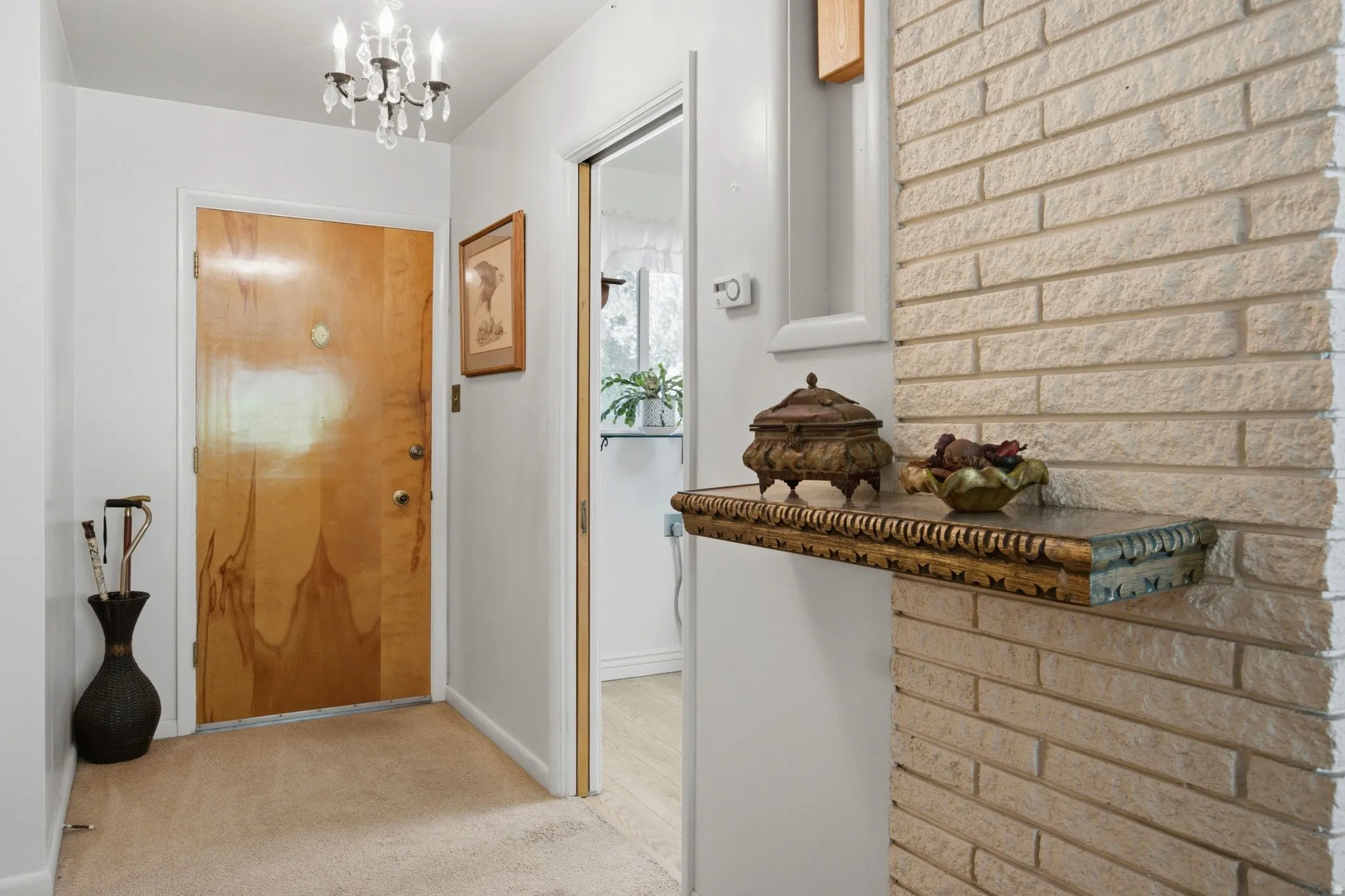 Natural wood-finish entry door with a crystal-accented chandelier, carpeted flooring, and a stone-clad wall with an integrated decorative shelf