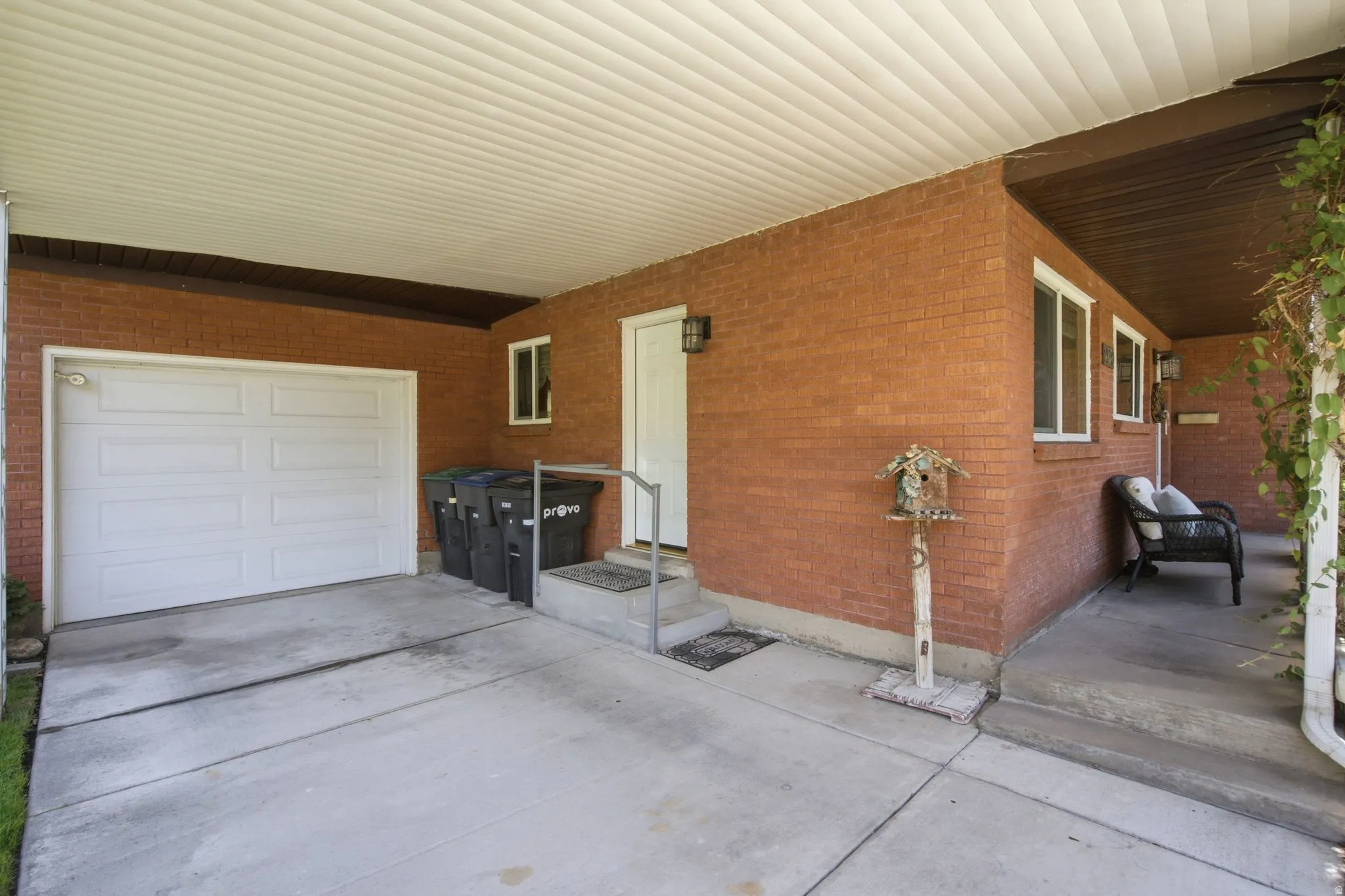 Spacious covered carport with polycarbonate roof offering weather protection, adjoining brick exterior with multiple entry points, concrete driveway and patio areas, and white overhead garage door providing secure vehicle storage