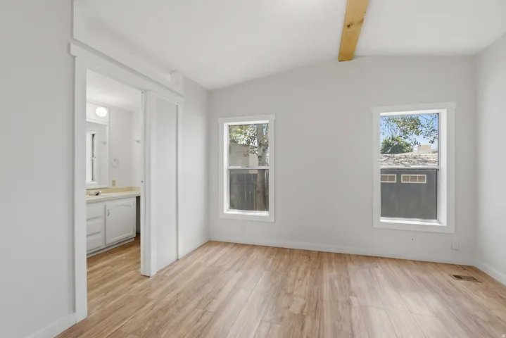 Vaulted ceiling with exposed wood beam, wood-finish flooring, two large windows, white walls, and a sliding barn door entry to a bathroom featuring a vanity with white cabinetry and a light-colored countertop