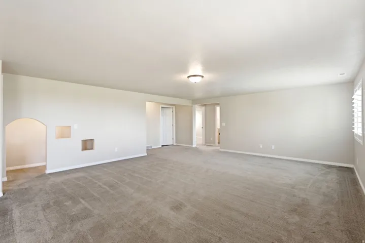Expansive carpeted room featuring an arched doorway, built-in wall niches, and window with white plantation shutters