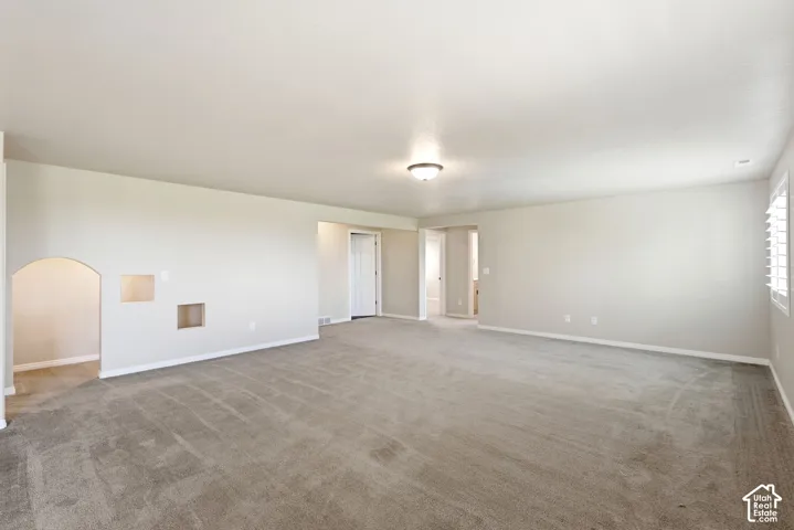 Expansive carpeted room featuring an arched doorway, built-in wall niches, and window with white plantation shutters