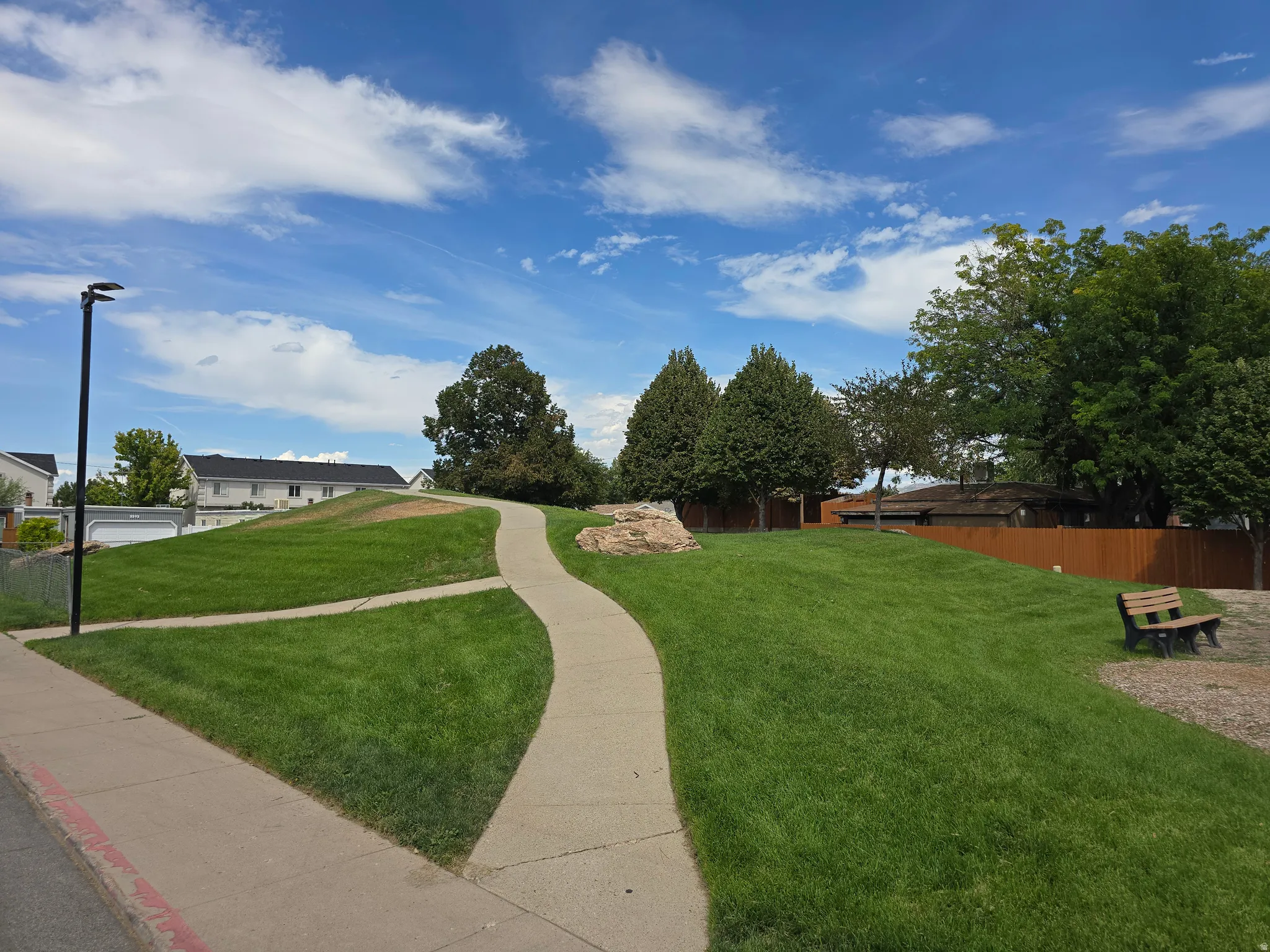 Expansive green space featuring a winding concrete pathway, mature trees, and a wooden bench