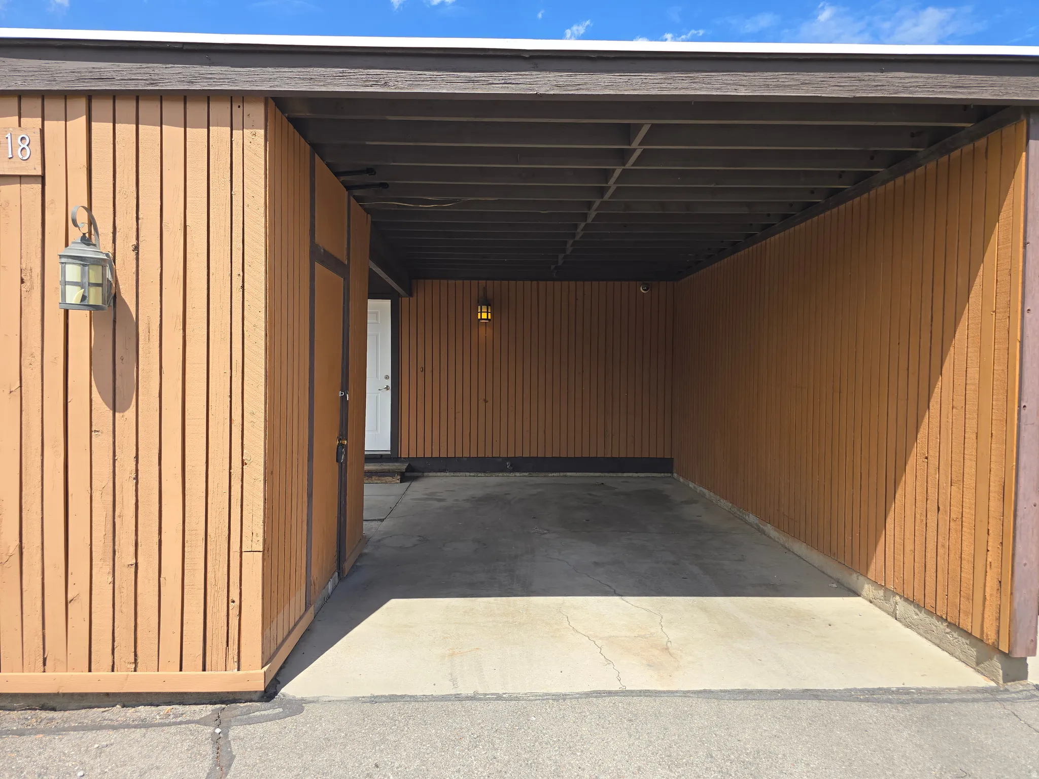 Carport entry with vertical wood paneling and concrete flooring, featuring an exterior wall sconce