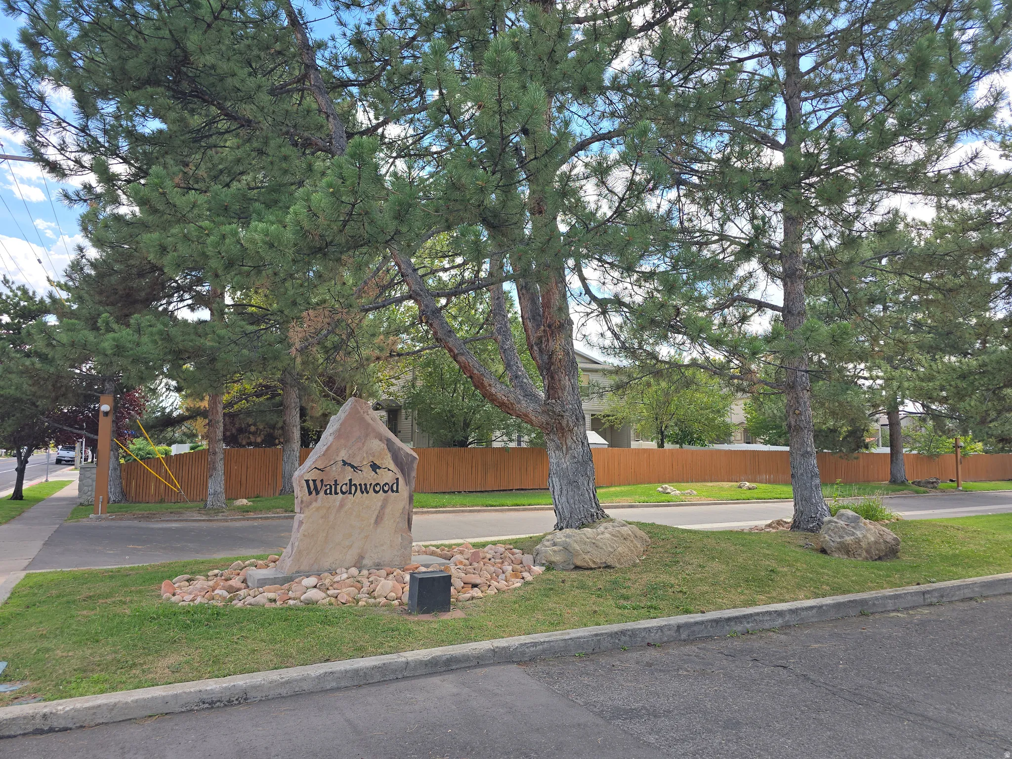 Stone community sign with decorative rock landscaping, mature trees, and a wooden perimeter fence