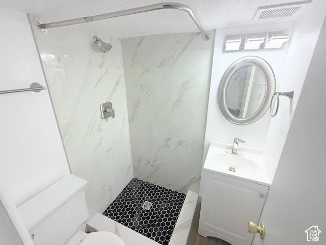 Walk-in shower with stone-finish tile surround, black hexagonal floor tile, pedestal vanity, oval wall-mounted mirror, and polished metal fixtures