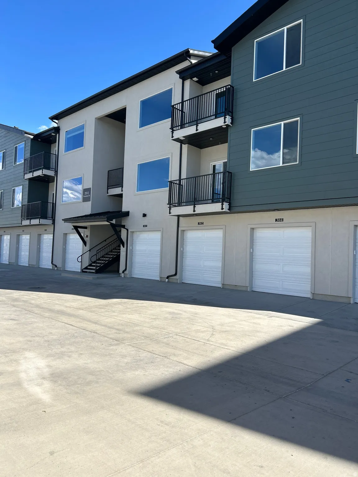 Multi-story building exterior featuring balconies with black railings, a mix of stucco and siding finishes, multiple windows, and ground-level garage doors