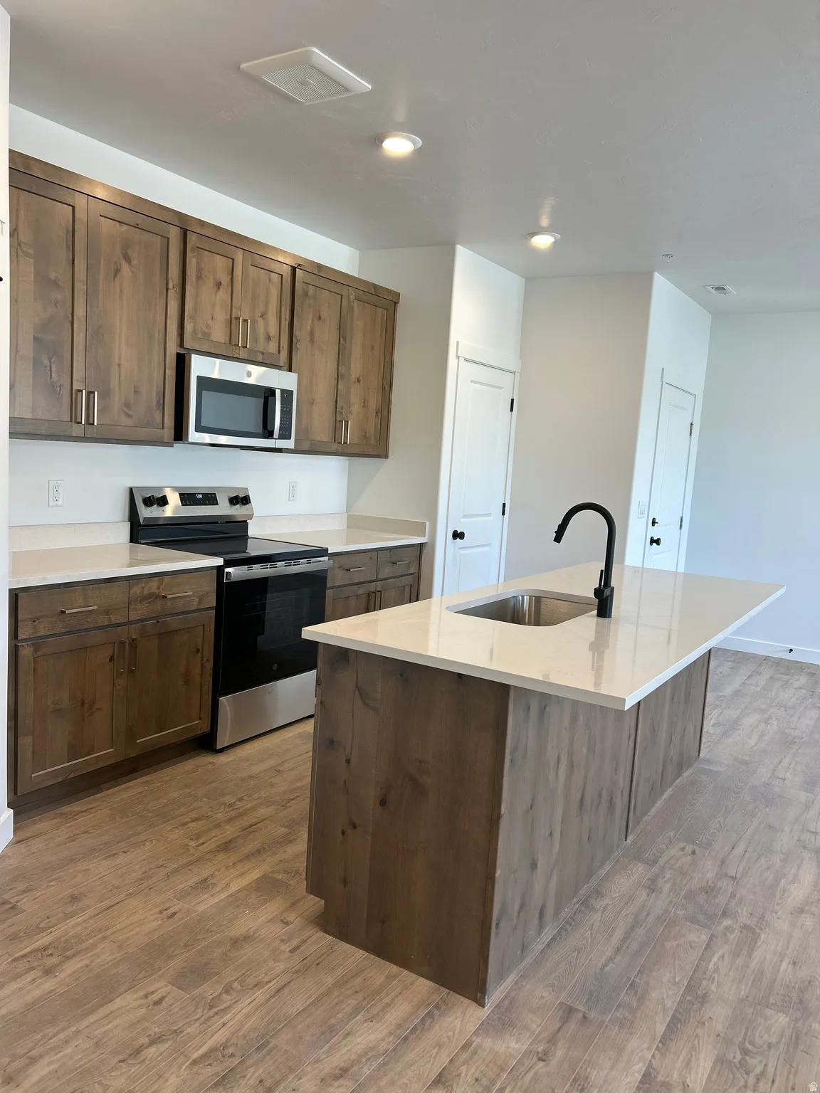 Kitchen featuring wood-finish cabinetry, stainless steel appliances, light-toned countertops, a center island with an undermount sink, and wood-finish flooring