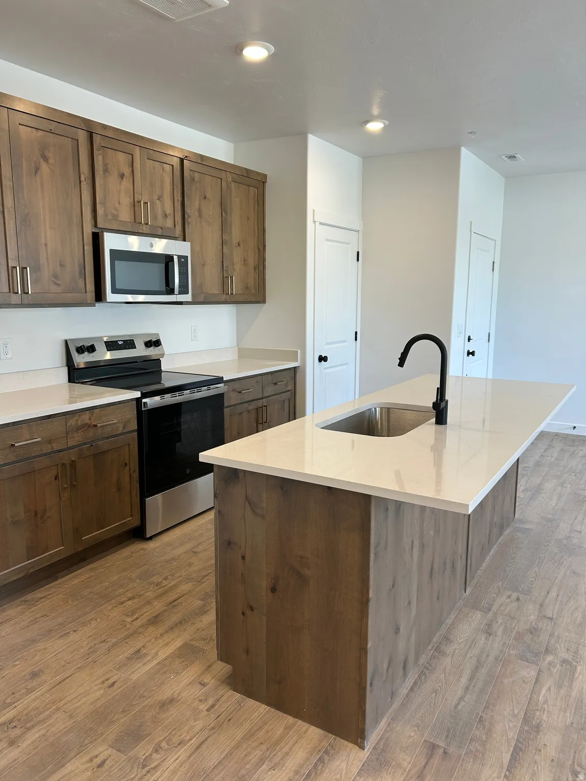 Kitchen featuring wood-finish cabinetry, light-toned countertops, stainless steel appliances, a center island with an integrated sink, and wood-finish flooring
