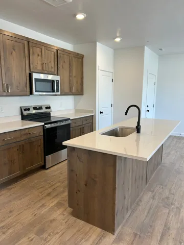Kitchen featuring wood-finish cabinetry, light-toned countertops, stainless steel appliances, a center island with an integrated sink, and wood-finish flooring