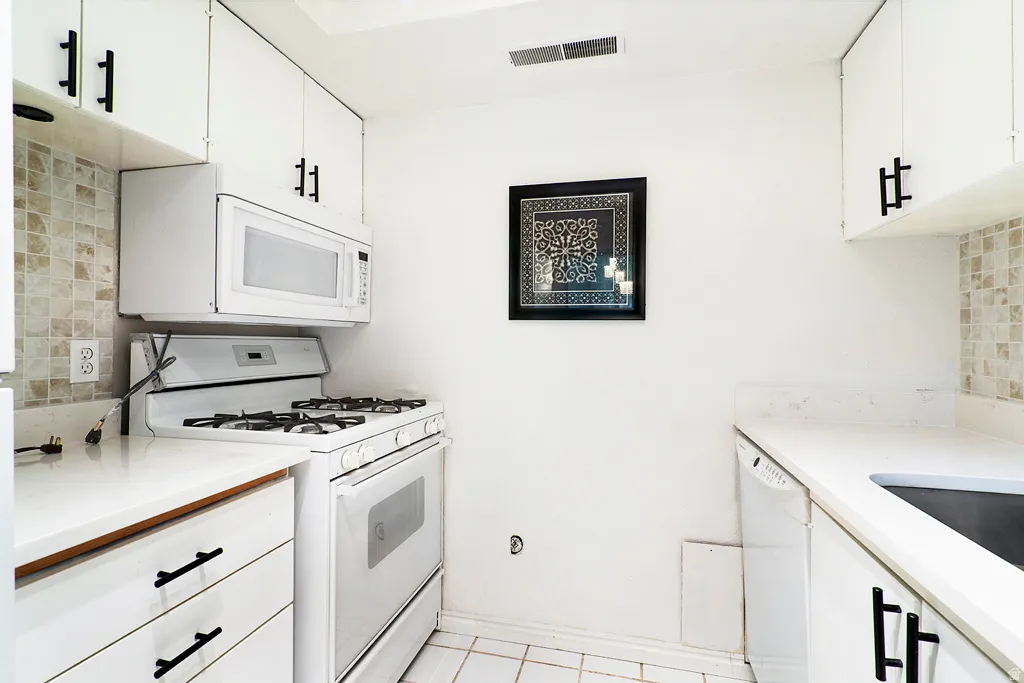 White cabinetry with black hardware, mosaic tile backsplash, white countertops, built-in microwave, and tile flooring
