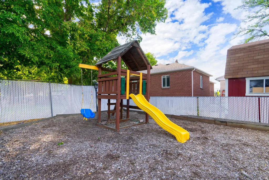 Mulch ground cover, white privacy fencing, mature trees, and a wooden play structure with a slide