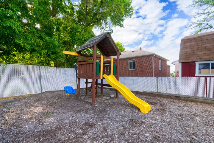 Mulch ground cover, white privacy fencing, mature trees, and a wooden play structure with a slide