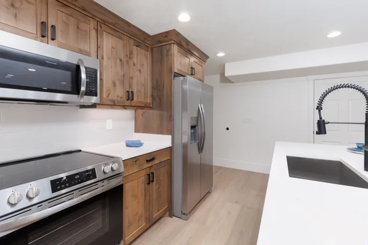 Basement  Kitchen Stainless steel appliances, wood-finish cabinetry, white countertops, white subway tile backsplash, and light wood-finish flooring