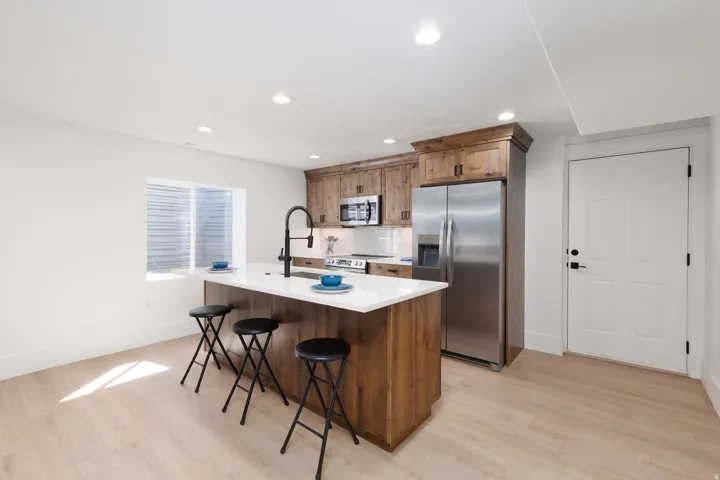 Basement Kitchen featuring a central island with white countertop, integrated sink with a matte black faucet, wood-finish cabinetry, and stainless steel appliances