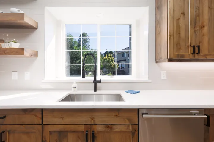 White framed window with grid inserts, stainless steel sink, black gooseneck faucet, light wood-finish cabinetry, and stainless steel built-in dishwasher