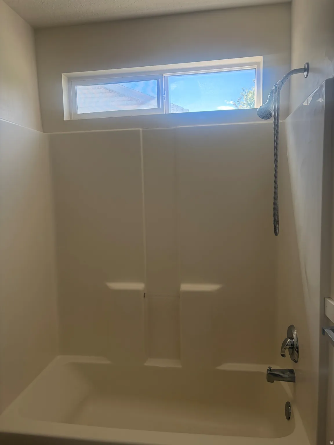Bathtub with integrated shower, chrome fixtures, and a high-set horizontal window