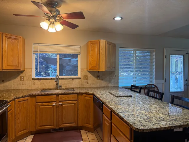 Kitchen featuring wood cabinetry, granite-finish countertops, tile backsplash, stainless steel sink, and a multi-light ceiling fan
