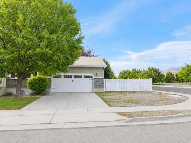 Two-car garage with white panel door, light-toned siding with stone-finish wainscoting, white privacy fence, concrete driveway, and mature trees