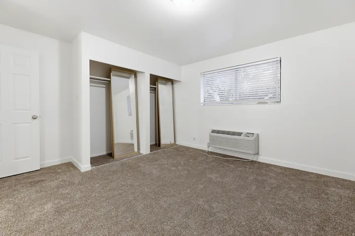 Plush carpeting, bright white walls, window with blinds, two mirrored closet doors, and a wall-mounted climate control unit