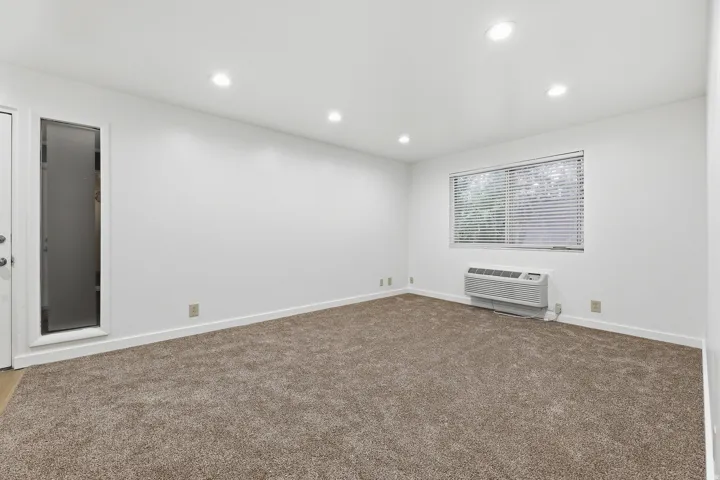 Recessed ceiling lighting, wall-mounted air conditioning unit, window with blinds, and neutral tone carpet