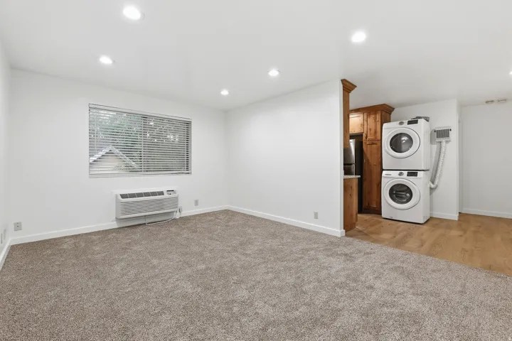 Recessed lighting, window with blinds, wall-mounted air conditioning unit, stacked laundry appliances, and wood-finish flooring