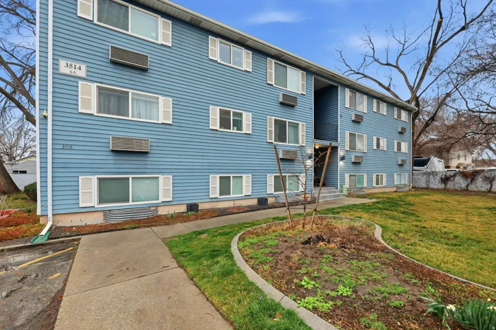 Modern multi-unit apartment building featuring light blue horizontal siding, expansive ground-level landscaping with curved planting beds and mature trees, and numerous windows providing natural light throughout the facade